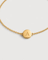 Macro detail of the Amara gold initial A medallion and fine chain on pale dove-grey