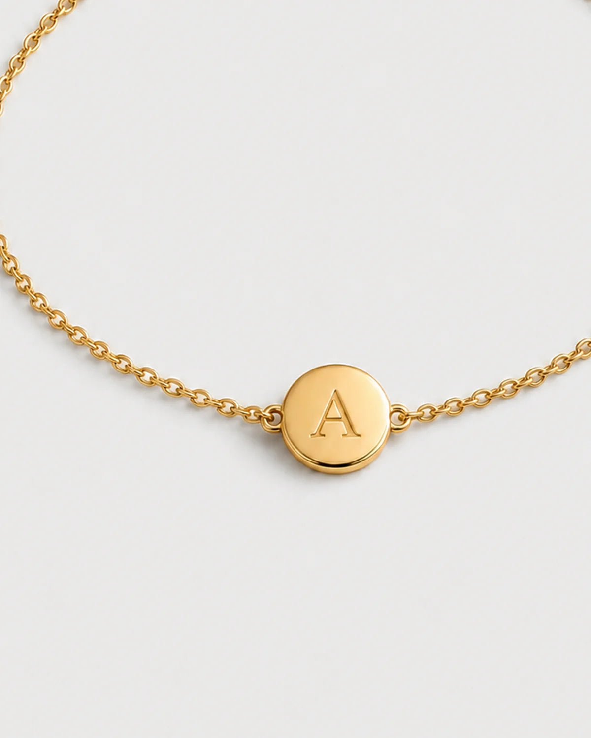 Macro detail of the Amara gold initial A medallion and fine chain on pale dove-grey