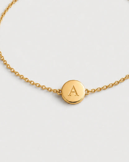Macro detail of the Amara gold initial A medallion and fine chain on pale dove-grey