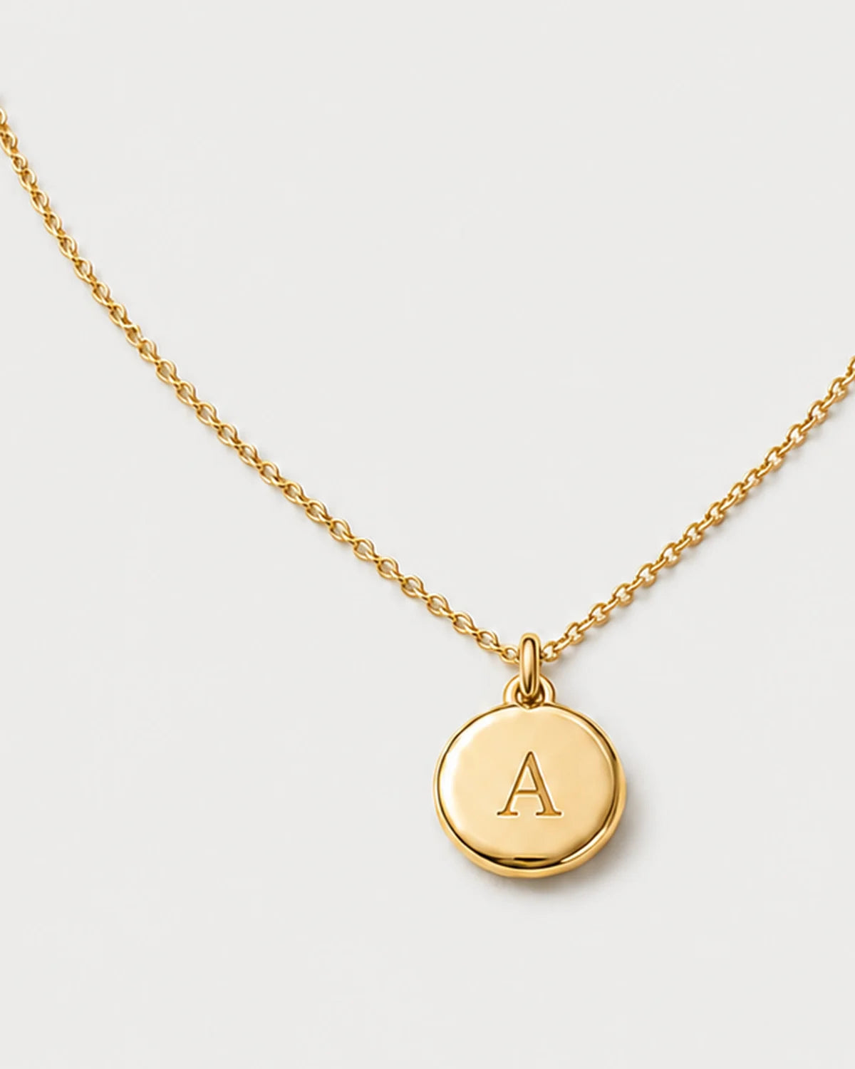 Macro detail of the Amara engraved A medallion and fine gold chain on pale dove-grey