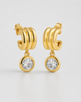 Pair of Astra gold triple-arc zircon drop earrings on pale neutral grey