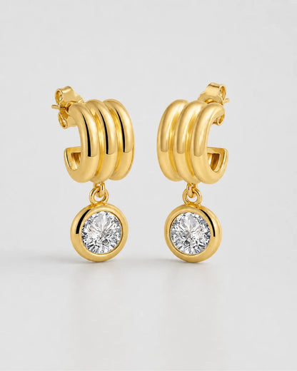 Pair of Astra gold triple-arc zircon drop earrings on pale neutral grey