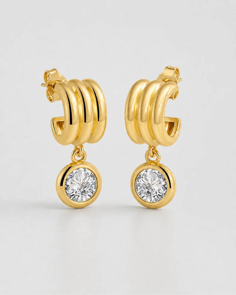 Pair of Astra gold triple-arc zircon drop earrings on pale neutral grey