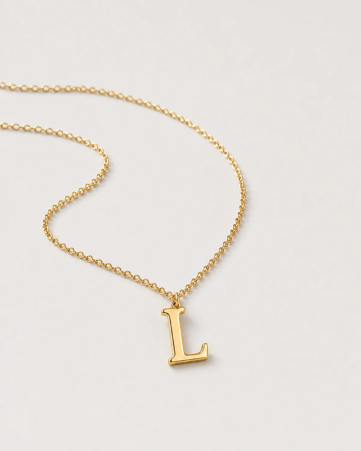 Macro view of the polished serif L charm and fine gold cable chain on pale dove-grey