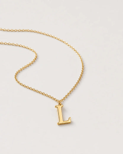 Macro view of the polished serif L charm and fine gold cable chain on pale dove-grey