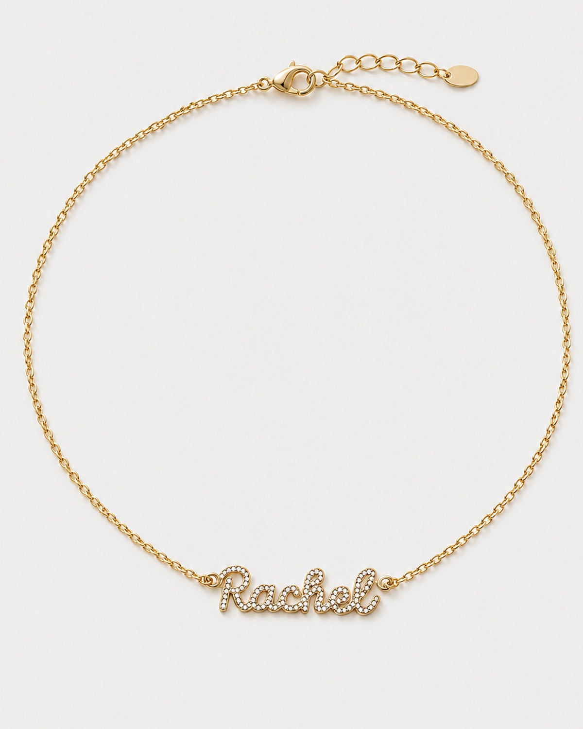 Mira gold pavé Rachel name necklace with round clasp, extender and oval tag on pale neutral grey