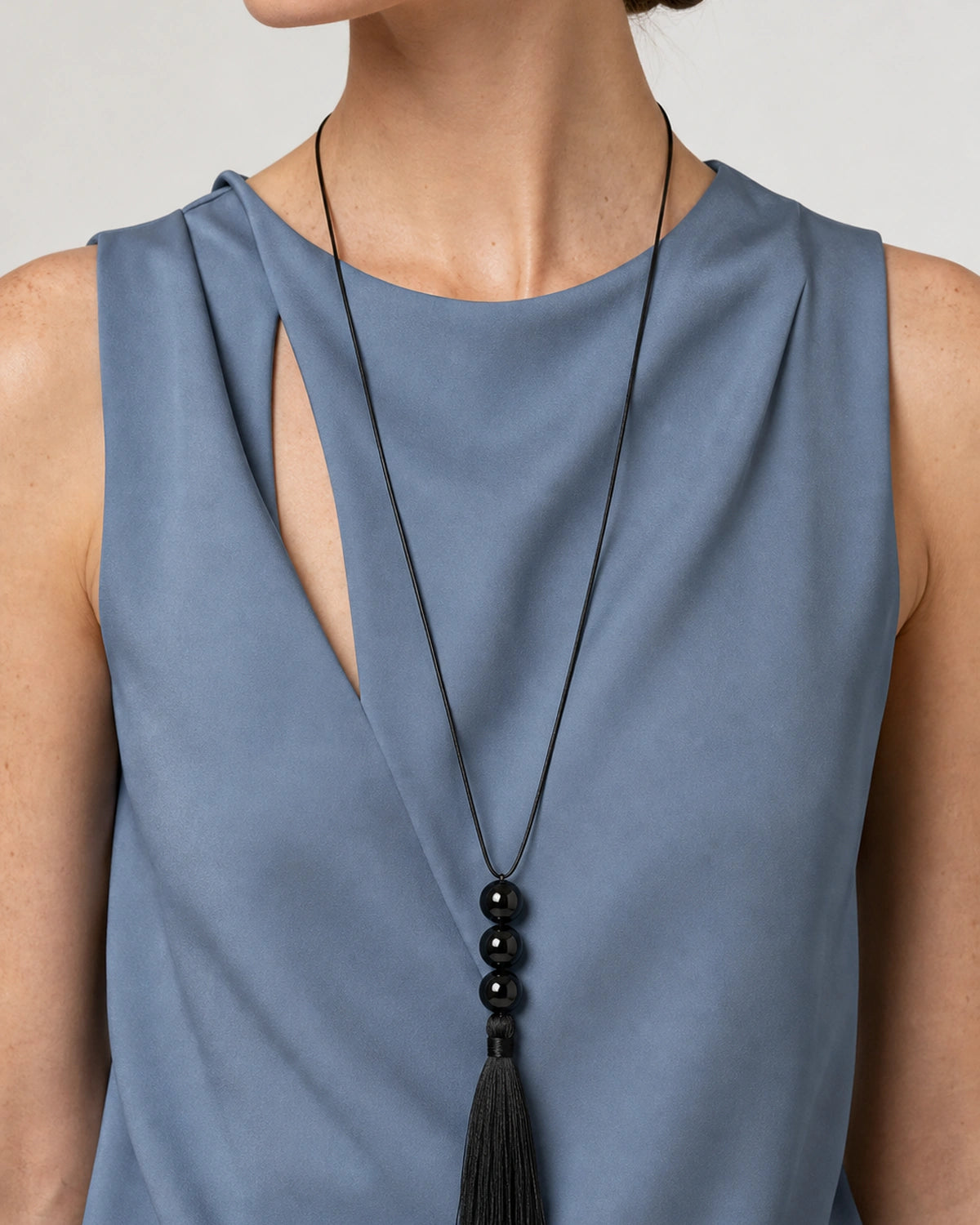 Mouth-down portrait wearing the Nera black tassel necklace with a mineral-blue draped top