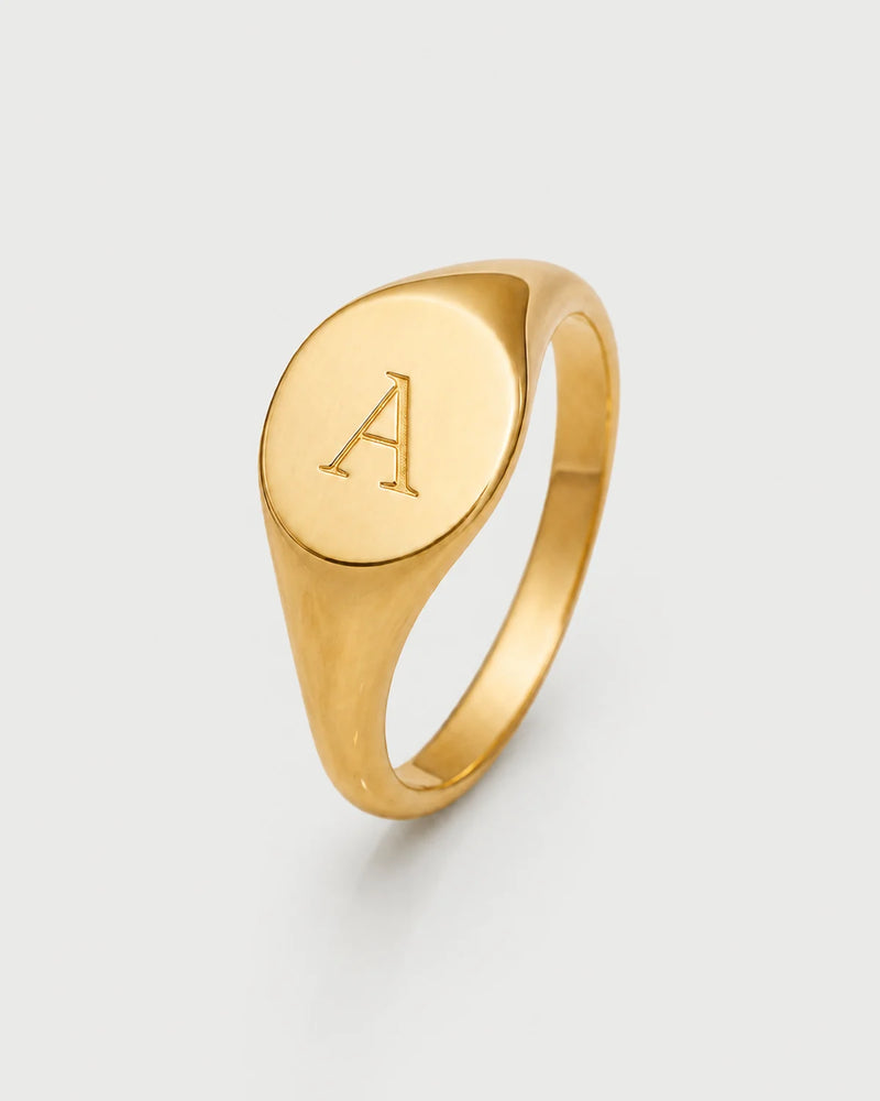 Solenne gold oval signet ring engraved with A on a seamless pale dove-grey studio background