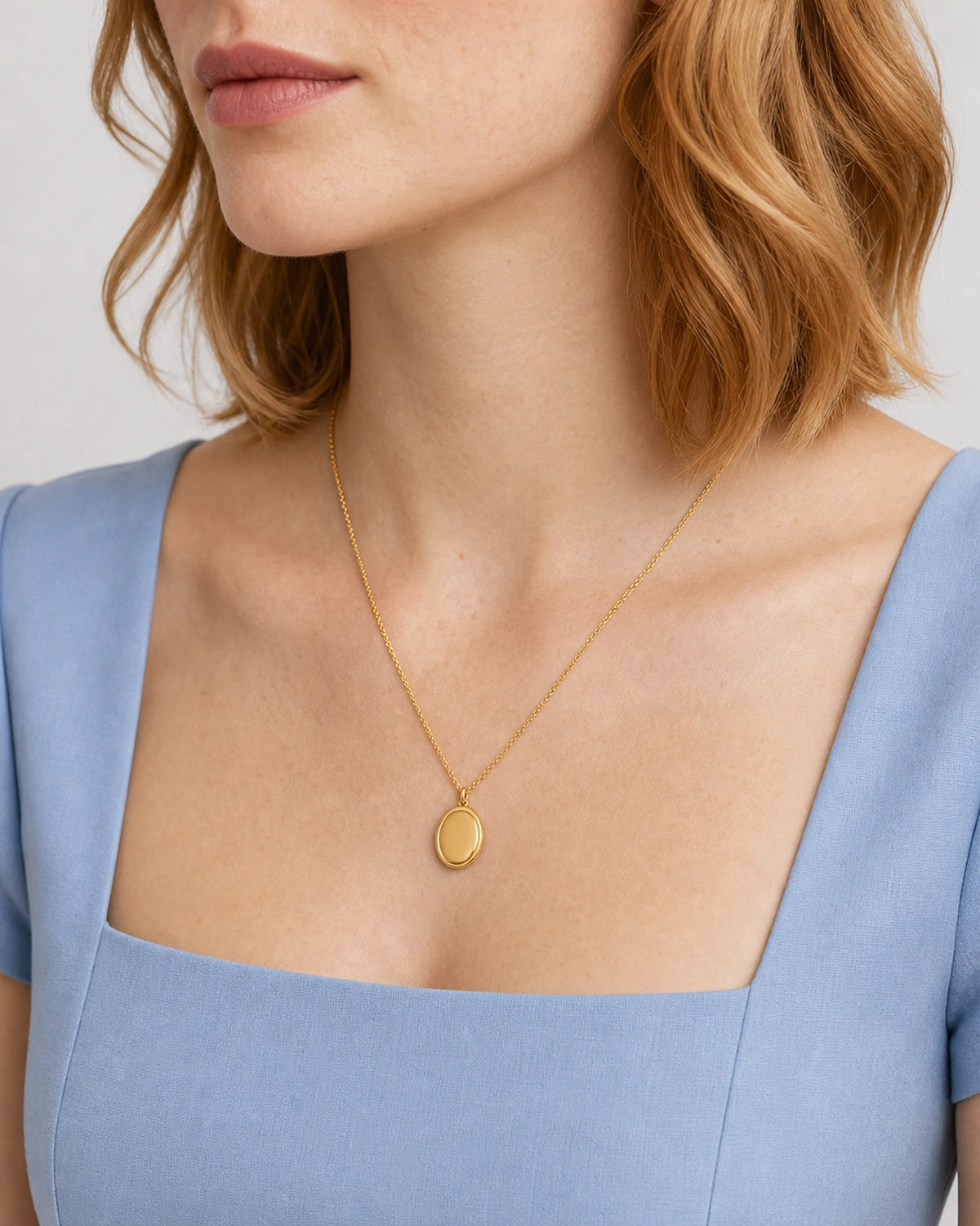 Mouth-and-chin-down portrait wearing the gold Solenne oval pendant with cornflower blue